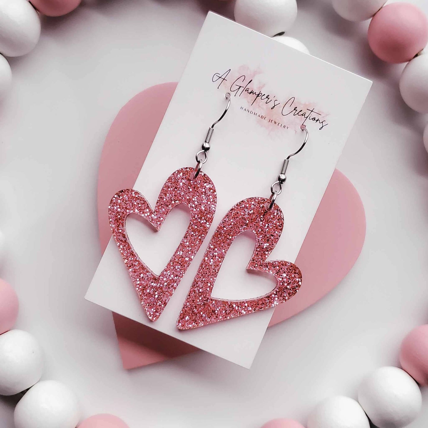 Heart earrings with the middle cut out in a heart shape as well. Made from pink glitter acrylic. Has silver hooks. The background is white with white and dusty pink beads.