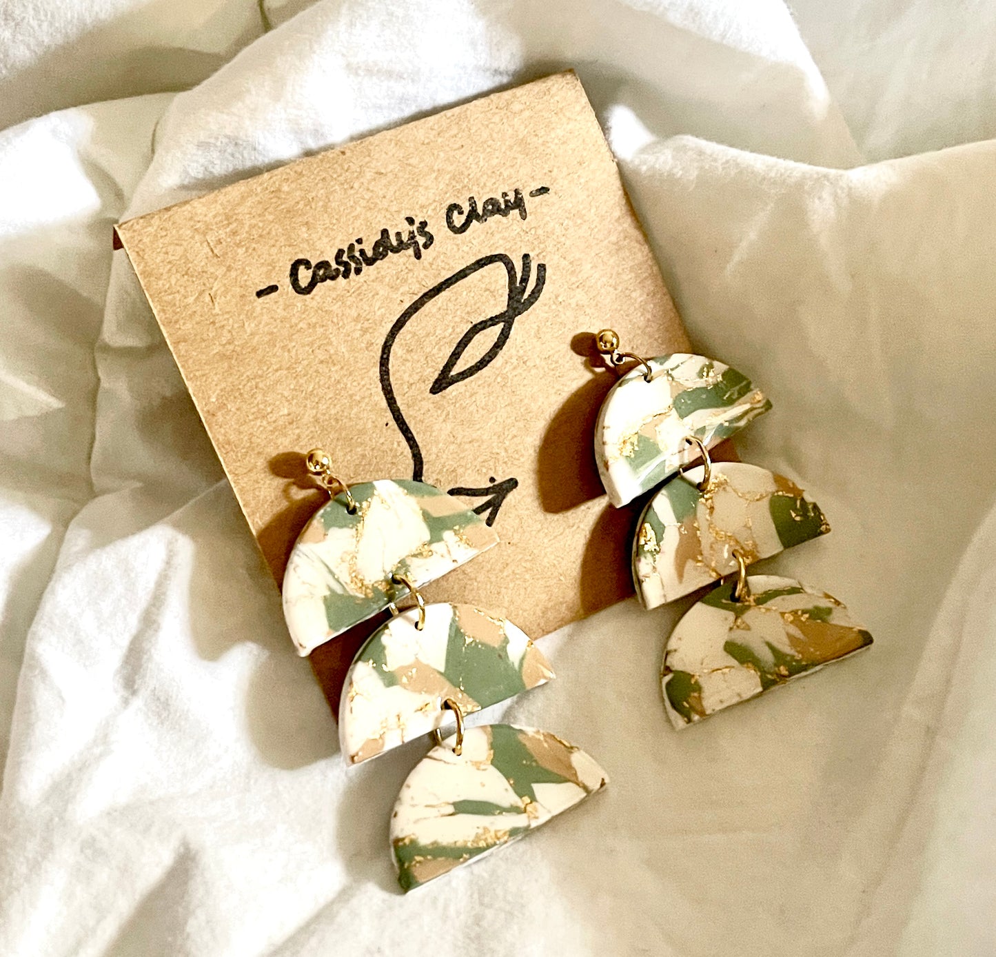 Olive green, cream, and gold marbled earrings. 3 tiers on half moon shapes held together by small gold rings. They are on a brown earring holder with white background.