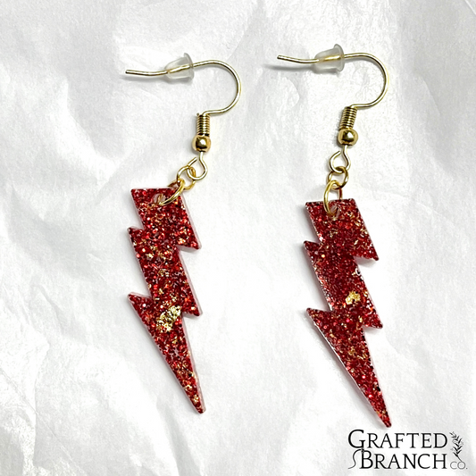 Earrings in the shape of a lightning bolt. They are red with gold flecks and gold hooks.