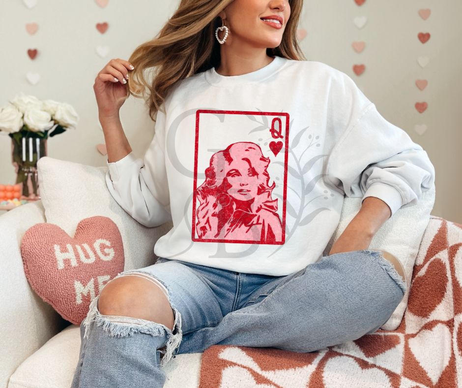 Woman sitting on a chair.  Valentine themed decor surrounding her. She is wearing a white sweatshirt with graphic of a playing card.  It is the queen of hearts with Dolly Parton on it.