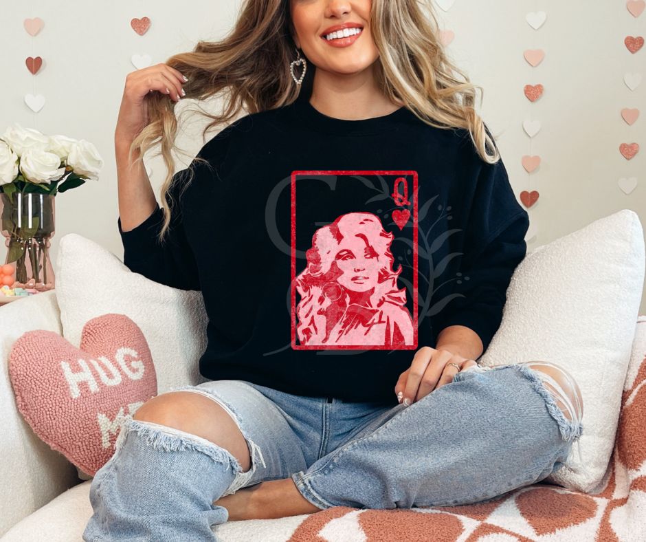Woman sitting on a couch.  Valentine themed decor surrounding her. She is wearing a black Sweatshirt with graphic of a playing card.  It is the queen of hearts with Dolly Parton on it.
