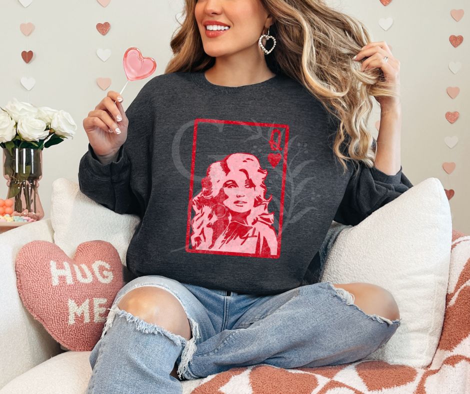 Woman sitting on a chair.  Valentine themed decor surrounding her. She is wearing a charcoal colored sweatshirt with graphic of a playing card.  It is the queen of hearts with Dolly Parton on it.