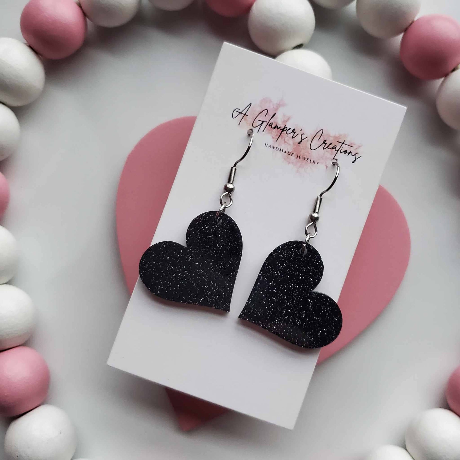 Black glitter heart earrings. Background is white and has white and dusty pink beads.