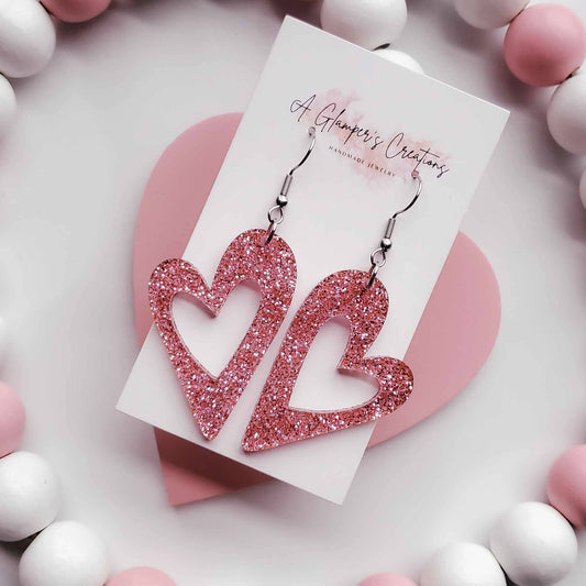 Heart earrings with the middle cut out in a heart shape as well. Made from pink glitter acrylic.  Has silver hooks.  The background is white with white and dusty pink beads.