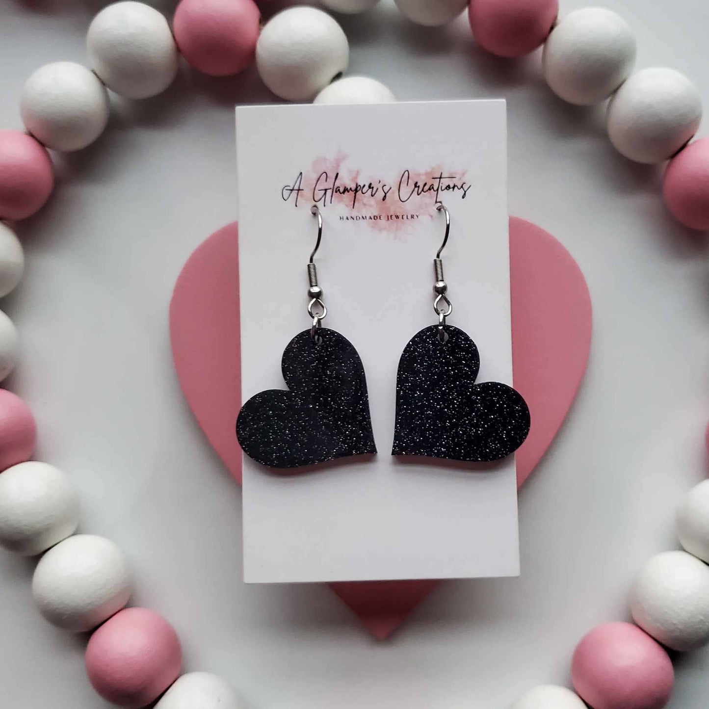 Black glitter heart earrings.  Background is white and has white and dusty pink beads.