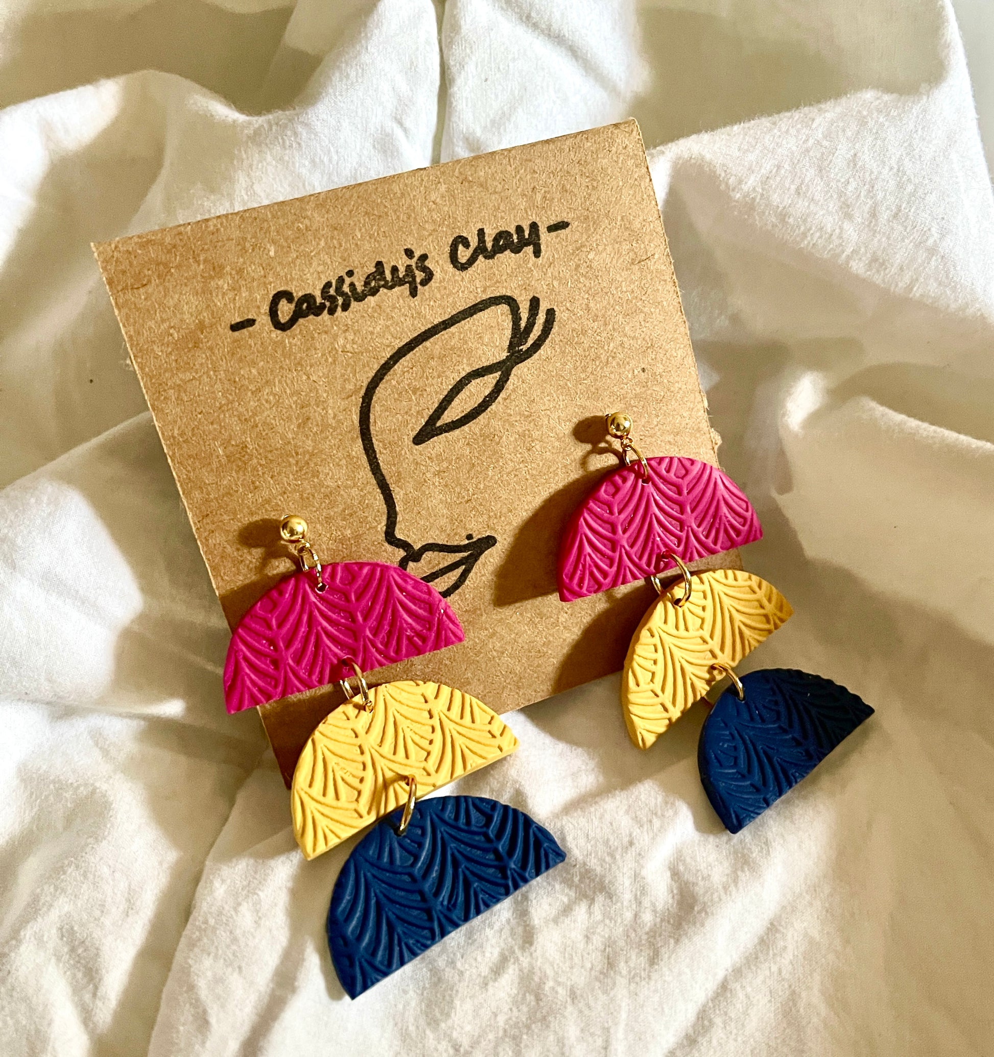 3 half moon shapes held together vertically by small gold rings.  First tier is pink, second tier is yellow, and third tier is navy.  They are on a cardboard earring holder. 2.5" long 1.5" wide