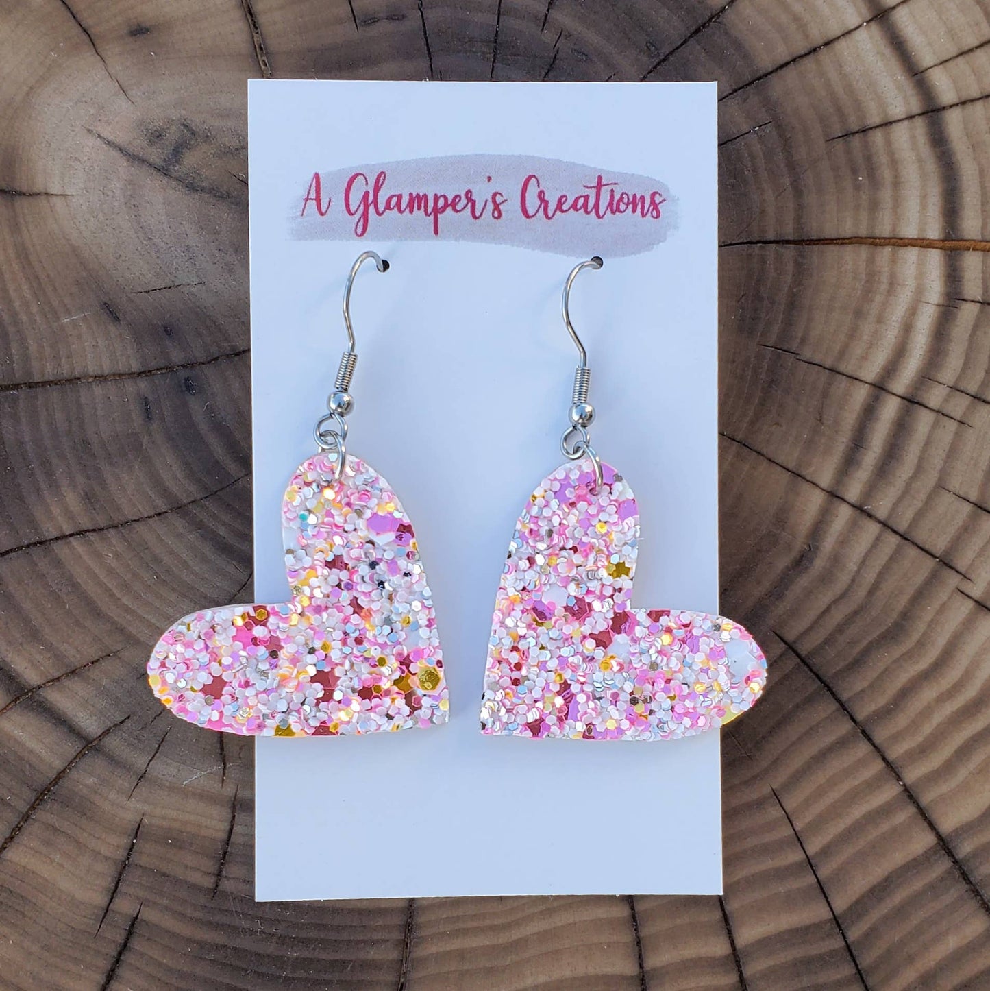 Light pink heart shaped leather earrings with thick, chunky glitter. Made with silver hooks.  Background is a natural wooden circle.